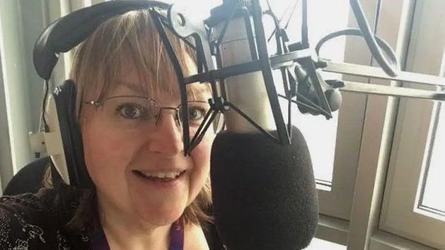 The late BBC Tees radio presenter, Julie Donaldson. She is wearing glasses, headphones and is talking into a large microphone.