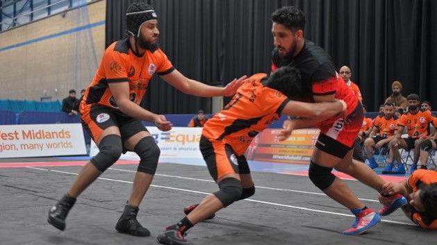 Kabaddi team Sandwell Kings players halt a raid during a British Kabaddi League match in a sports hall, as teammates look on from chairs behind them.