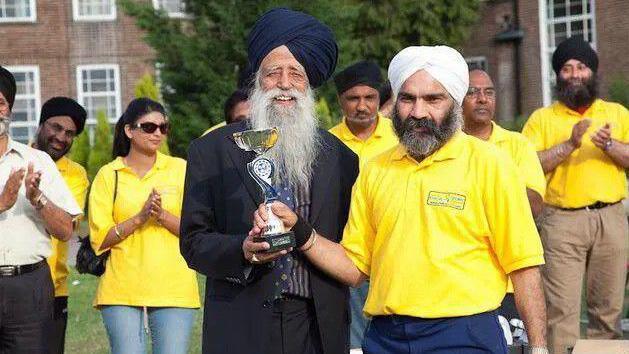 Fauja Singh on the left has a white beard and navy turban. He is smiling while wearing a black blazer and dark striped shirt. He is handing a trophy to Meva Singh Dhesi on the right who has a greying beard and a white turban. He wears a bright yellow polo shirt. People in the background are clapping while also sporting the yellow polo.