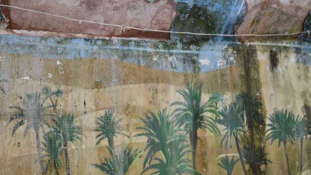 An old wall, looking dirty and damp with a fading and damaged mural of palm trees