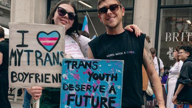 Two people, both wearing sunglasses, holding signs, one says "I love my trans boyfriend" and the other says "trans youth deserve a future"