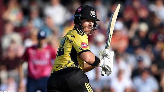 D'Arcy Short, wearing a black helmet, black trousers, a yellow shirt and white gloves, plays a shot while batting for Gloucestershire on 29 May 2026