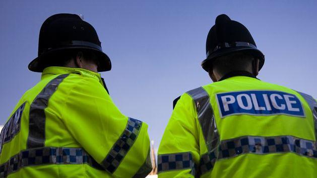 Two police officers in helmets and hi-vis jackets stand together with their backs to the camera.