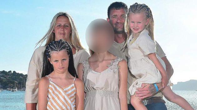 The family stands in front of the sea. Mr Roller is holding is daughter, who has blonde, braided hair, in one arm, his two other daughters stand next to him and his partner. The face of the daughter in the middle is blurred to protect her identity.