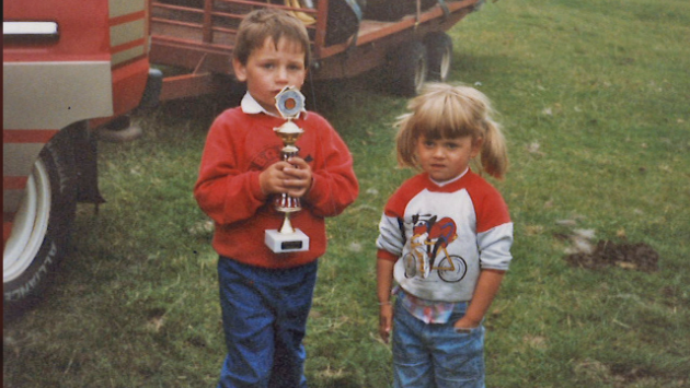 a young boy with brown hair and wearing a red sweatshirt and jeans holds a sporting trophy in his hands. A young girl with blonde hair in bunches, wearing a red and white sweatshirt and jeans stands next to him in a field where part of a truck and trailer can be seen in the background