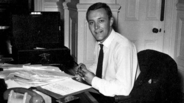 A black and white image of Tony Benn sitting in his office in front of his desk with a pile of papers in front of him