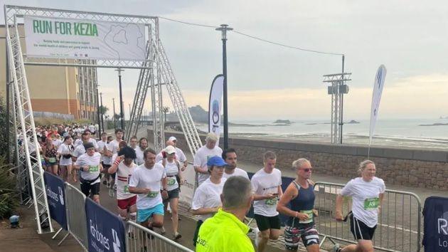 Most of the runners are wearing white tops with a green square with their race numbers on. Some are in fancy dress. They run under a banner saying Run for Kezia. In the background to the left is a hotel building and then behind the sea wall next to the runners - the beach stretches out into the distance.