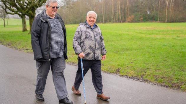 Two men walking along a path next to grass, one with a walking stick