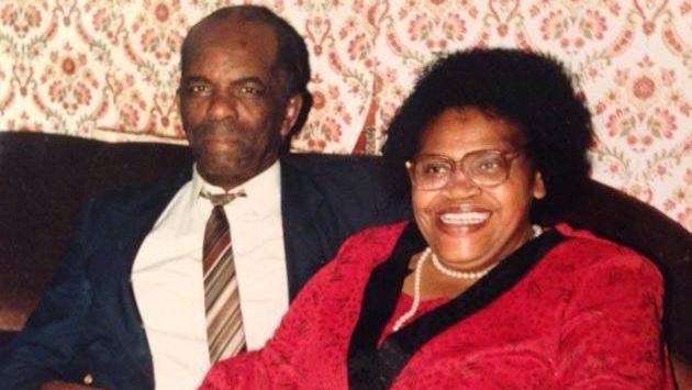 Ms Baton's parents sitting on a brown chair, and Mr Banton has his arm around Ms Burrell. He is wearing a blue suit jacket, white shirt and striped tie. He has very short black hair. Ms Burrell is wearing a red jacket and white pearl necklace, and has short black hair.