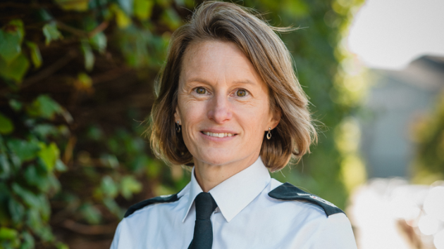 A woman with shoulder length brown hair stands against a background of greenery and looks at the camera. She's wearing a police uniform white short-sleeved shirt with epaulettes and a dark tie with emblem.