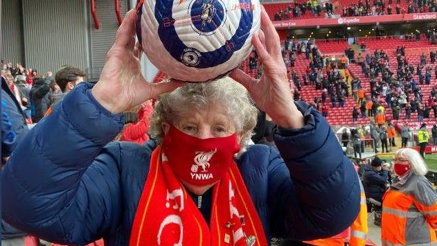 Peggy Pickavant is holding the match ball following a game at Anfield. She has raised the ball above her head and is standing in the Kop. She is wearing a Liverpool FC mask across her face and has a scarf around her neck.
Credit: Peggy Pickavant