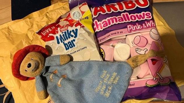 A Paddington bear comforter on a postal package beside a white and red packet of Milky bar buttons and a pink and purple packet for haribo marshmallows.