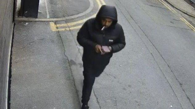 Rehman, dressed in black with his hood up, walking along a pavement - as caught on CCTV.