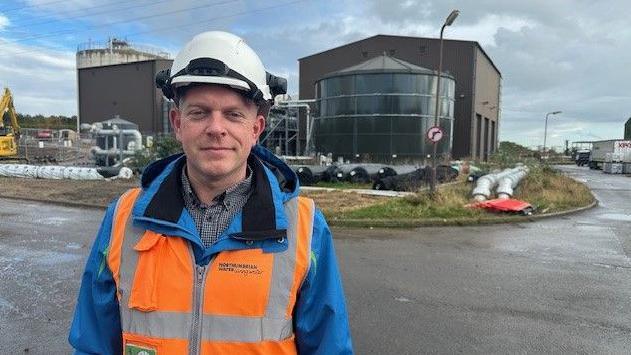 Tony Rutherford, the treatment works manager at Northumbrian Water's Howdon plant. He is wearing an orange and blue high viz jacket and a hard hat. Behind him can be seen a number of black and silver pipes on the ground and two industrial buildings connected by a series of pipes.