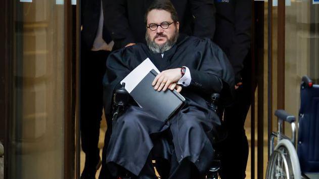 Nicholas Rossi in a wheelchair being pushed out of Edinburgh High Court, He is wearing a black robe, glasses and his a grey beard.