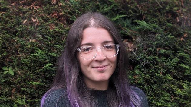 woman with dyed purple hair, glasses and facial piercings standing in front of a hedge or tree