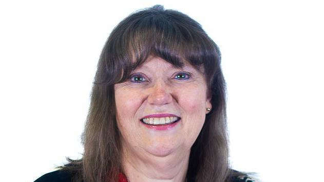 Cate Caruth is smiling at the camera in front of a white background. She has shoulder length brown hair with a fringe. She is wearing a dark coloured top.