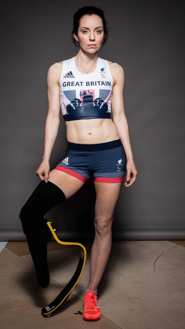A full-body shot of Stef Reid in her GB Paralympic kit. She is white, has brown hair and her running blade is clearly visible