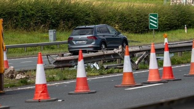 A dual carriage way with a black car in the distance and a row of orange cones in the forefront. The barrier in the middle of the road is badly damaged.