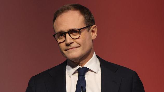 Tom Tugendhat wears black glasses and a dark suit with a white shirt and navy tie in front of a red background. 