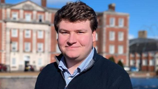 A head and shoulders picture of Roger Quilliam standing outside with a large four-storey red brick building blurred in the background. It is a bright sunny day and the sky is blue. He is wearing a dark blue zip up jumper over a light blue striped shirt which is open at the neck.