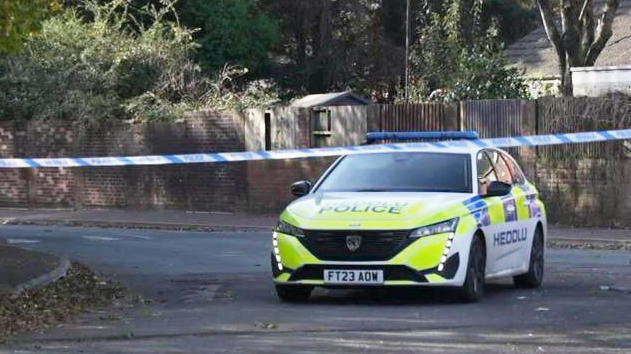 A police car and a police tape stretched across the road, Heol Trostre, in St Mellons, Cardiff. A house is in the background, and trees and fencing