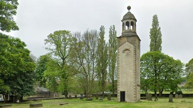 A church tower in a green grassy graveyard surrounded by trees