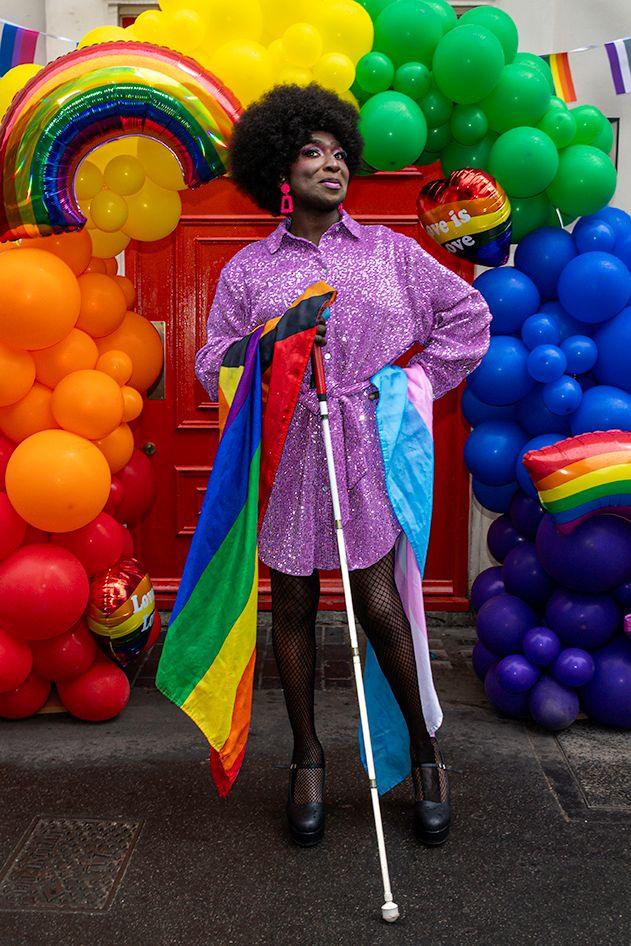 A drag artist with black afro hair, wearing a pink sparkly dress and large bright pink earrings. They are holding a white cane and a rainbow flag, and posing with the other hand on their hip, over which is draped a trandgender pride flag. Behind them is a multicoloured balloon arch and a rainbow helium balloon.
