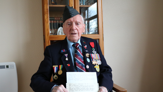 An elderly man in a blue suit with lots of military medals pinned to his jacket. He  wears a blue military cap and holds a telegram in his hands. He is sitting in a chair in the room of a house, with a cabinet behind him and a radiator to the left.