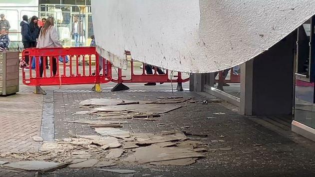 A large white stretch of plasterboard has fallen from a row of shops, and there is debris all over the ground.