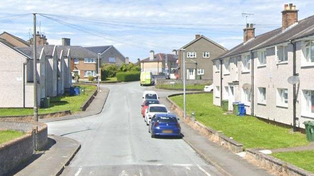 Four cars and a van can be seen parked on the road, with cream and light brown fronted houses to either side of the road.