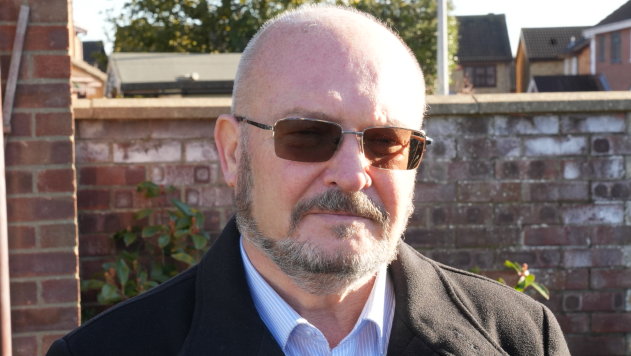 Graham Plant is wearing dark tinted sunglasses and is looking at the camera. He is standing in front of a brick wall and is wearing a striped blue shirt and black coat. 