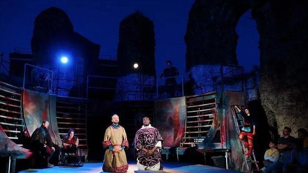 A performance in the ruins of Reading Abbey with bright theatre lights shining down on two actors kneeling centre stage and others stage left and right looking towards them.  