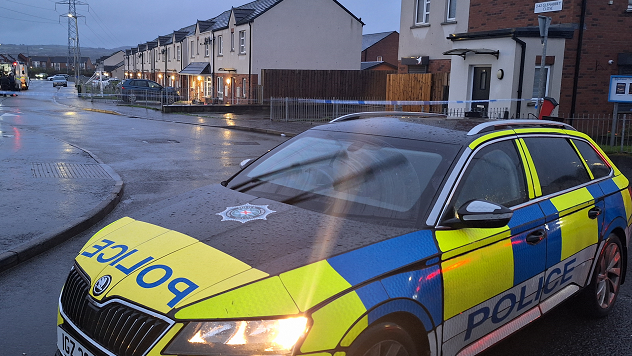 A police car sits in front of a row of houses. It is early morning and the sky is dark. a police tape can be seen behind the vehicle