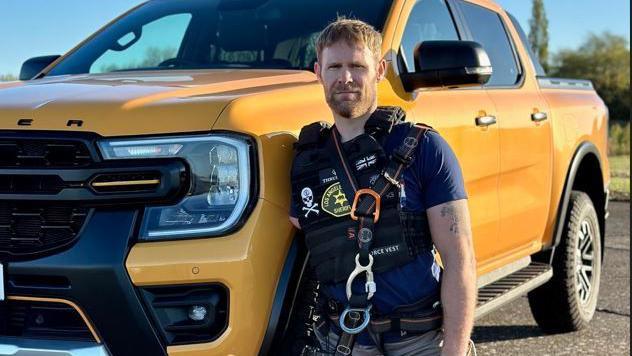 Mark Ormrod MBE stands to the right of a large yellow vehicle, looking directly at the camera. There are various attachments on his torso, used to connect to the vehicle when he pulls it 