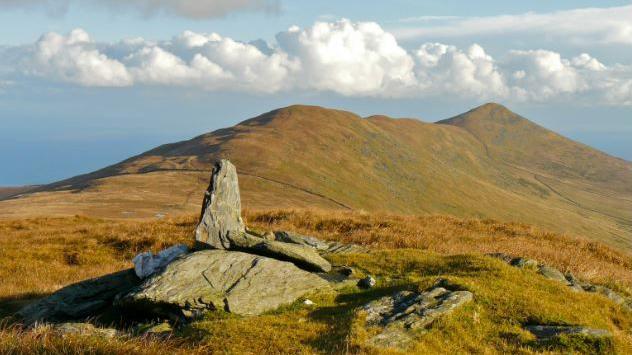 Rolling green hills, with some orange coloured patched. Some large stones can been seen in the foreground. The sky is blue and has white fluffy clouds.