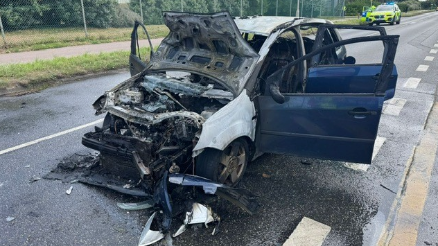 A car which has been ablaze - the front of the bonnet is collapsed on the ground with all the doors open. Police car in the background of the road.