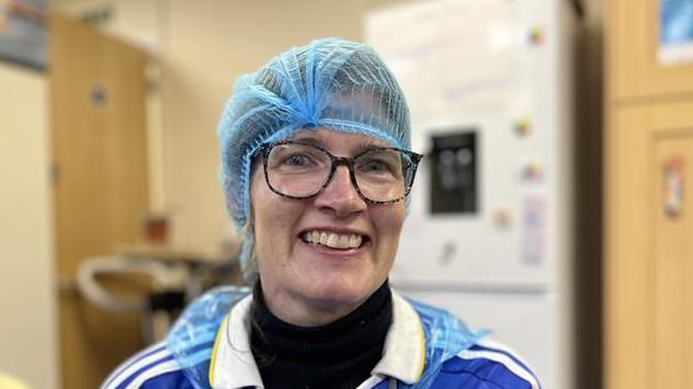 A woman smiles at the camera. She has a blue hair net over her head and she is wearing glasses along with a blue shirt which has white stripes over the shoulders. 