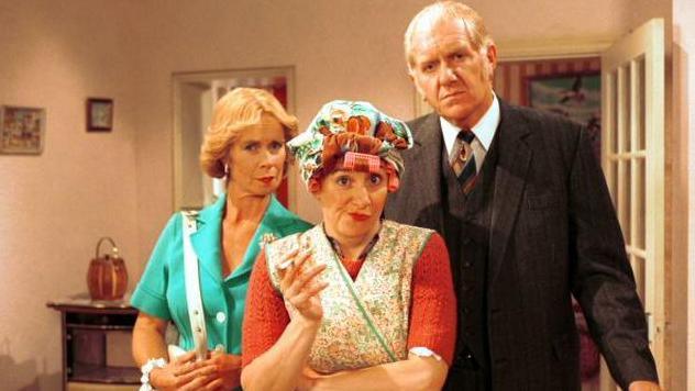 Actors Celia Imrie, Victoria Wood and Duncan Preston pose in Victoria Wood's Sketch Show. Victoria Wood is in a cleaner/dinnerlady outfit. Celia Imrie is dressed in a turquoise dress and Duncan Preston wears a three-piece suit. The image is from 2001.