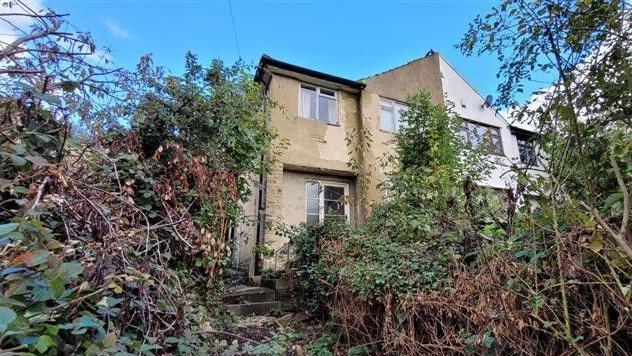 A beige end terrace house with steps leading up to it, surrounded by overgrown hedges. 