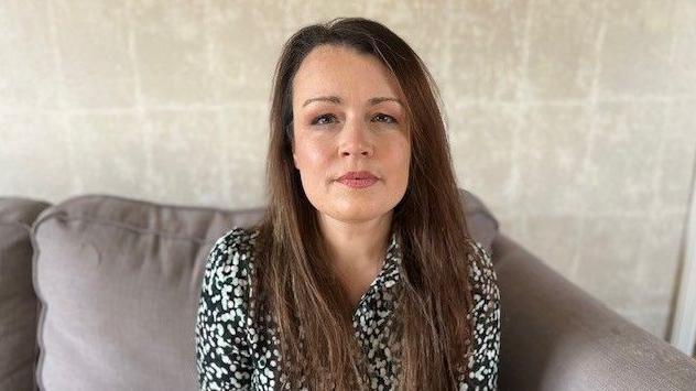 A woman with long dark hair, wearing a black and white top sitting on a sofa, staring at the camera