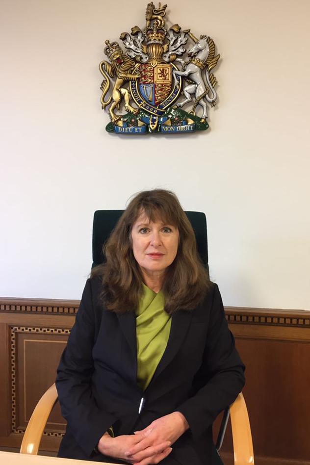 Senior coroner Jacqueline Lake wearing all black and sitting in a chair under a large royal crest