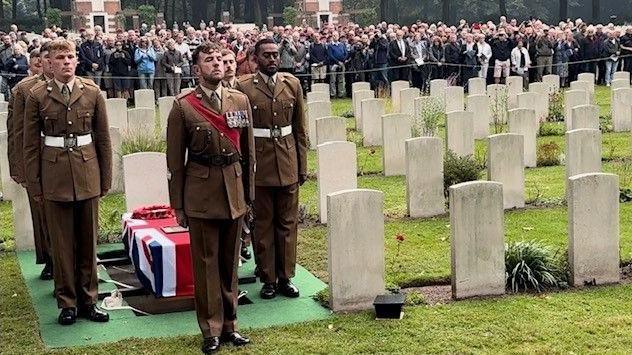 A commemoration service is held at Oosterbeek war graves cemetery, showing a group of soldiers standing to attention around a coffin wrapped in a union flag, with white gravestones to their right