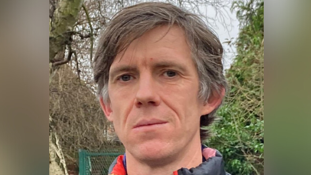 Ash Lever was stood in the middle of the image looking at the camera. He had short grey and brown hair and was wearing a coat- standing in front of hedges.
