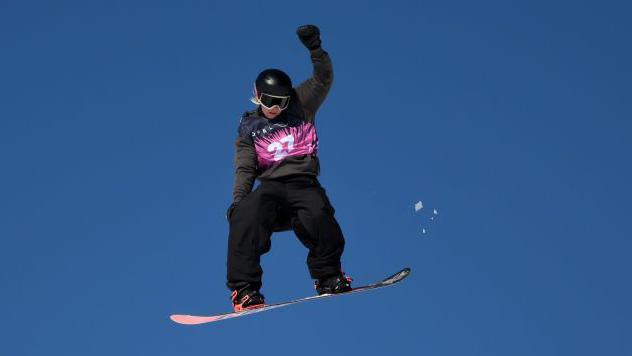 Maisie Hill flying through the air on a snowboard with one arm held up in the air during a competition in 2025