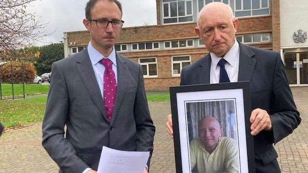 Amos Waldman, who has glasses and brown hair and wears a grey suit with a pink tie, and Dr Steven Waldman, look at the camera with sorrowful expressions while holding a framed photo of a smiling Dan Kay