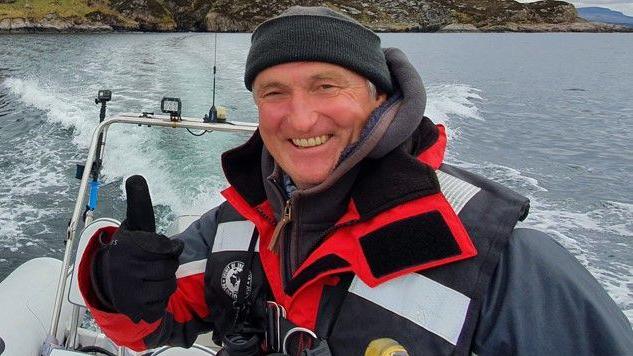 A man standing on a boat gives a thumbs up while smiling. He is wearing a black hat, black gloves and a red and blue coat. 