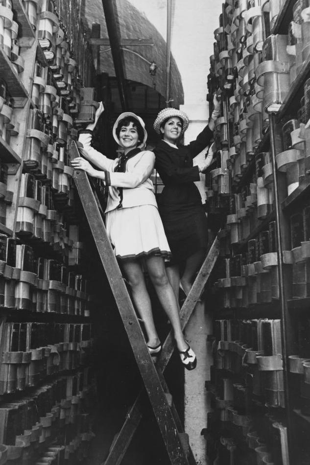 Two women at the Somerset House records office balance on ladders between the record books. Black and white archive image.