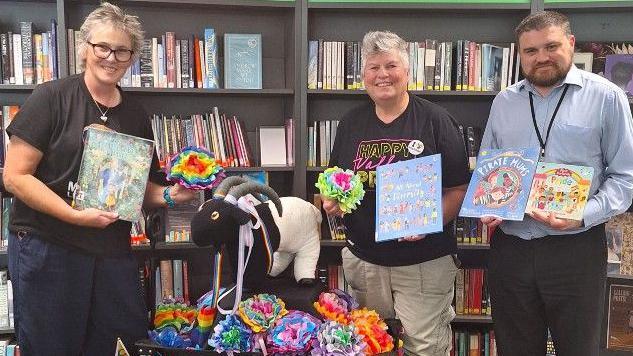 Three people stand in front of bookshelves, each holding up children's books about the LGBTQ+ community. In the middle of them is a mini parade float, which is black and features a ram. It is decorated with rainbow fabric and flowers