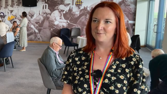 A woman with red hair, wearing a black floral dress and rainbow lanyard stands in front of the camera